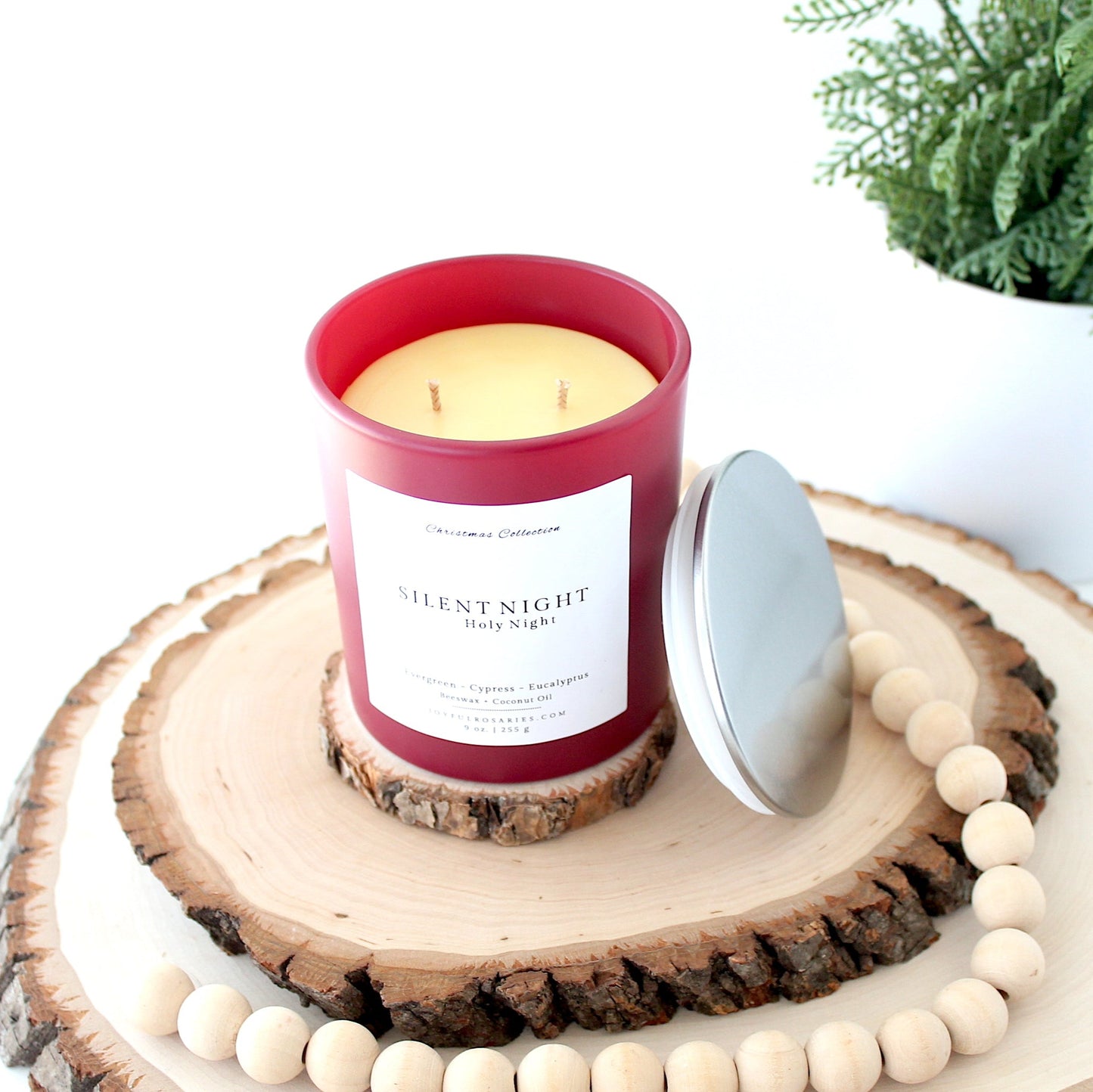 Candle in a red container and silver lid labeled 'Silent Night' on a wooden surface with a plant in the background.