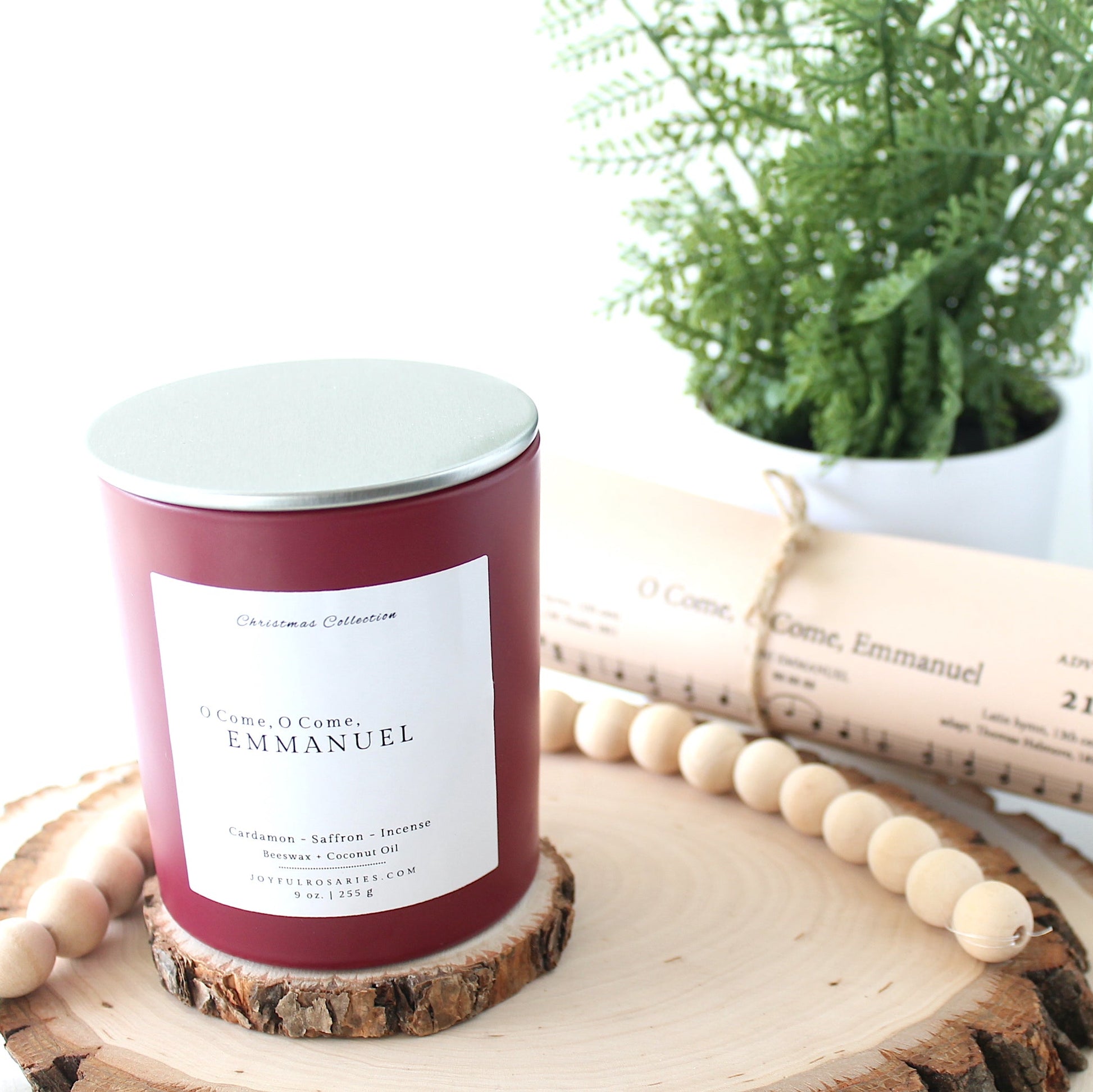 Red Candle with a silver lid and square label on a wooden surface next to a plant and rolled paper.