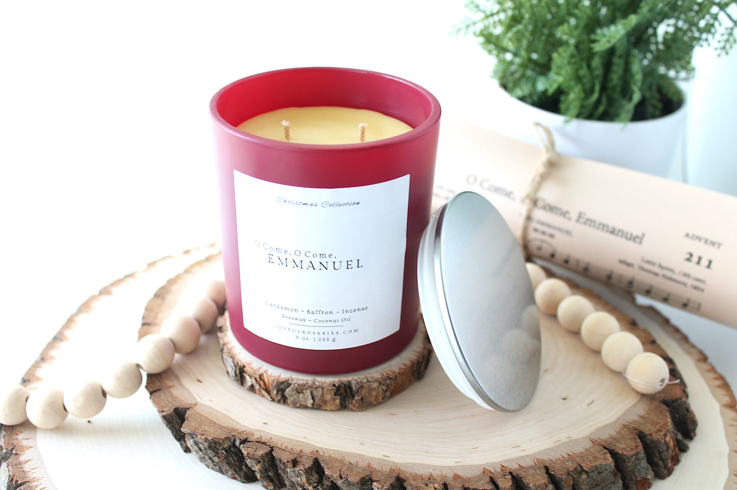 Candle with a red container and white label with tilted silver lid on a wooden coaster with a plant in the background