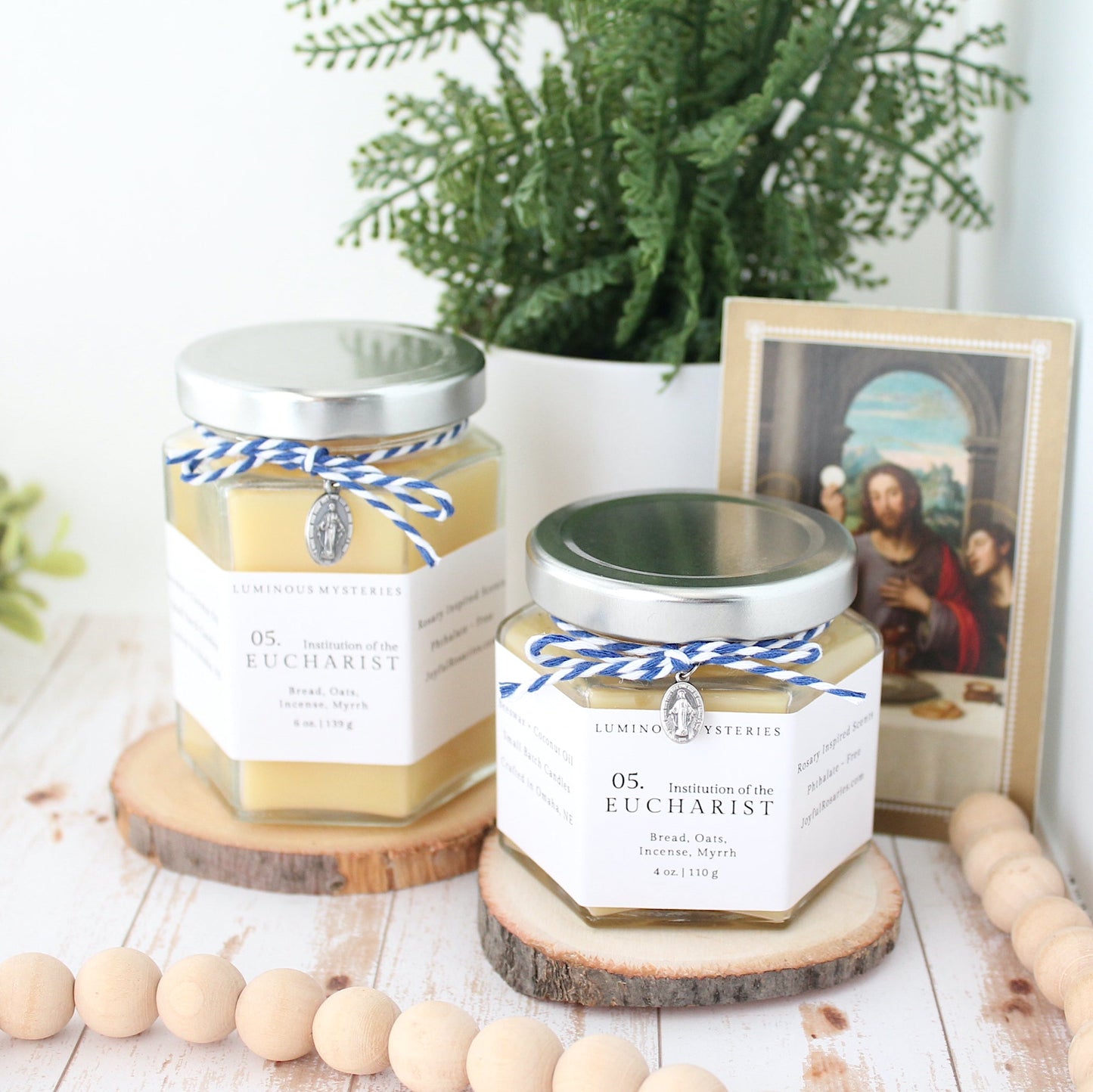 Two candles with labels on a wooden surface, surrounded by decorative items like a plant and beads.
