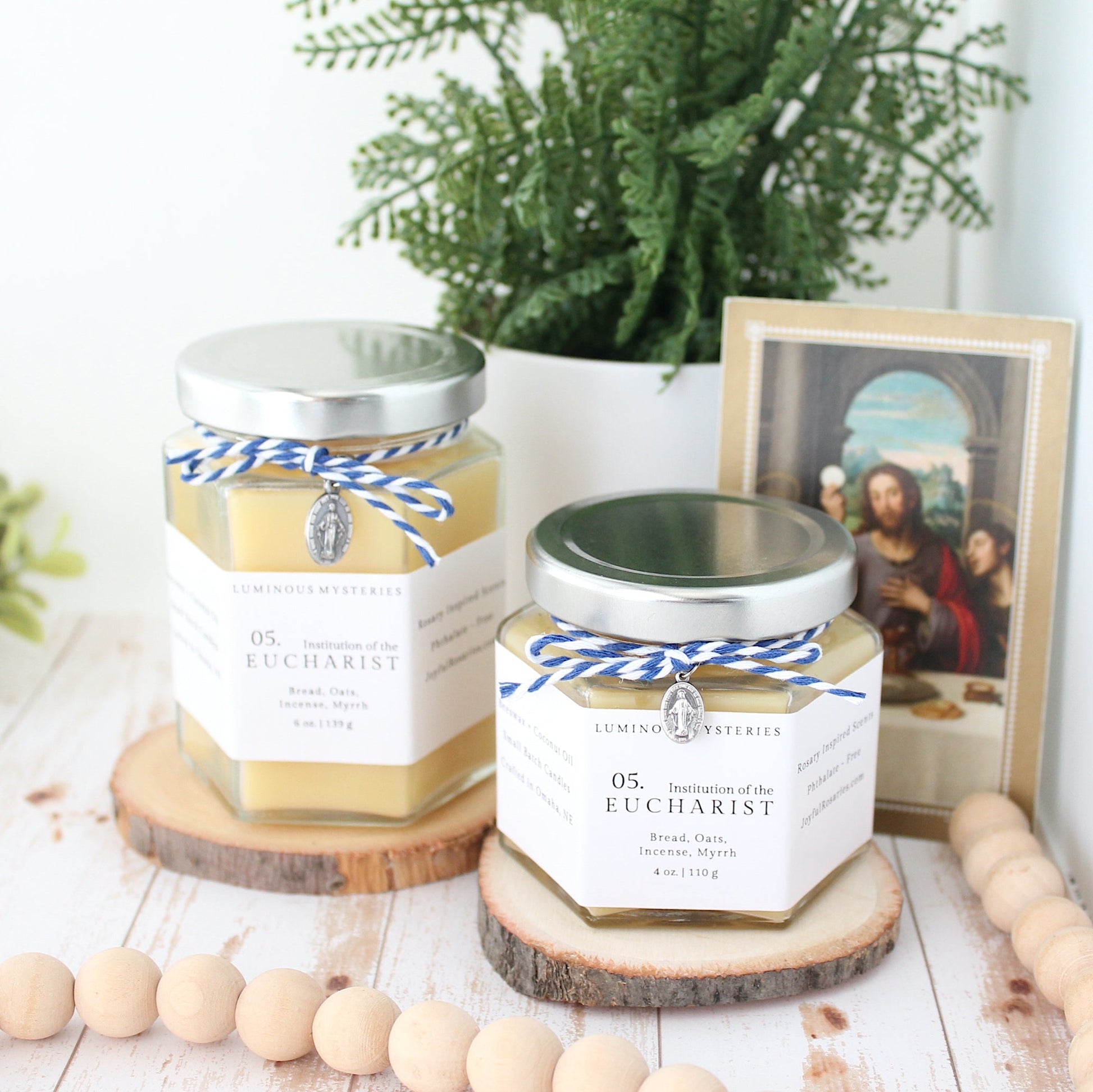 Two candles with labels on a wooden surface, surrounded by decorative items like a plant and beads.