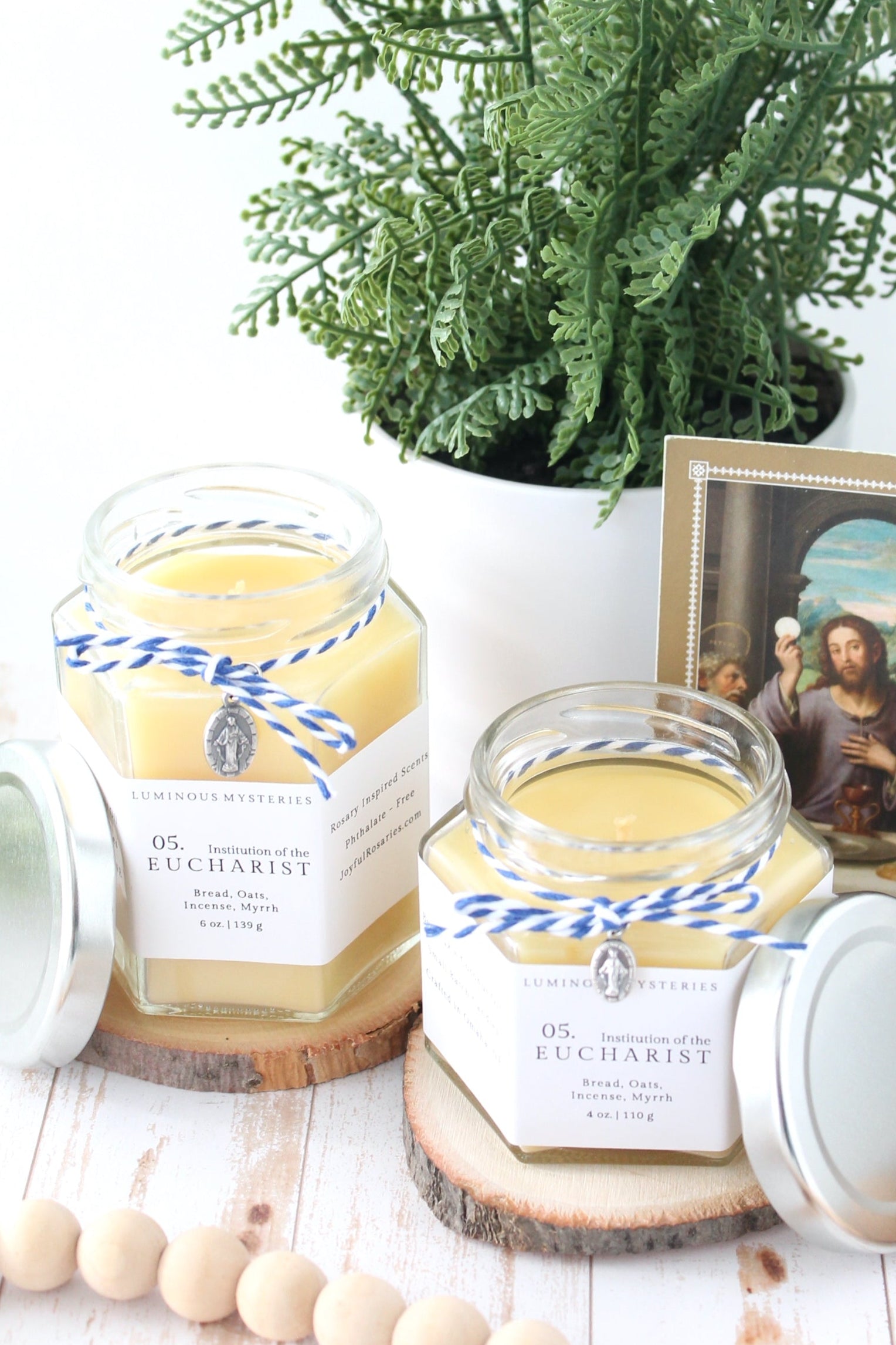 Two jars of 'eucharist' candles with decorative ribbons on a table with a plant and framed picture in the background.