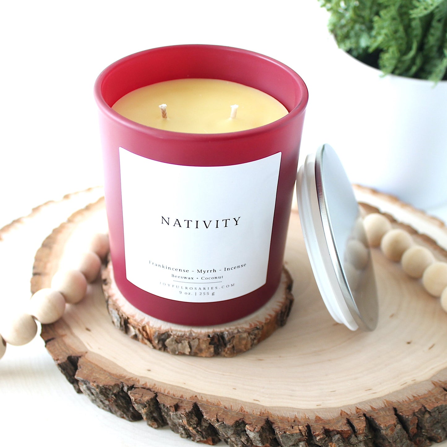 Candle in a 'Nativity' container on a wooden coaster with a plant in the background